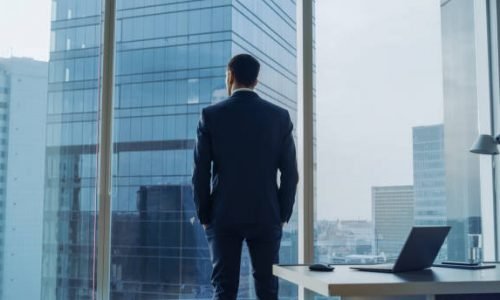 Back View of the Thoughtful Businessman wearing a Suit Standing in His Office, Hands in Pockets and Contemplating Next Big Business Deal, Looking out of the Window. Big City Business District Panoramic Window View.